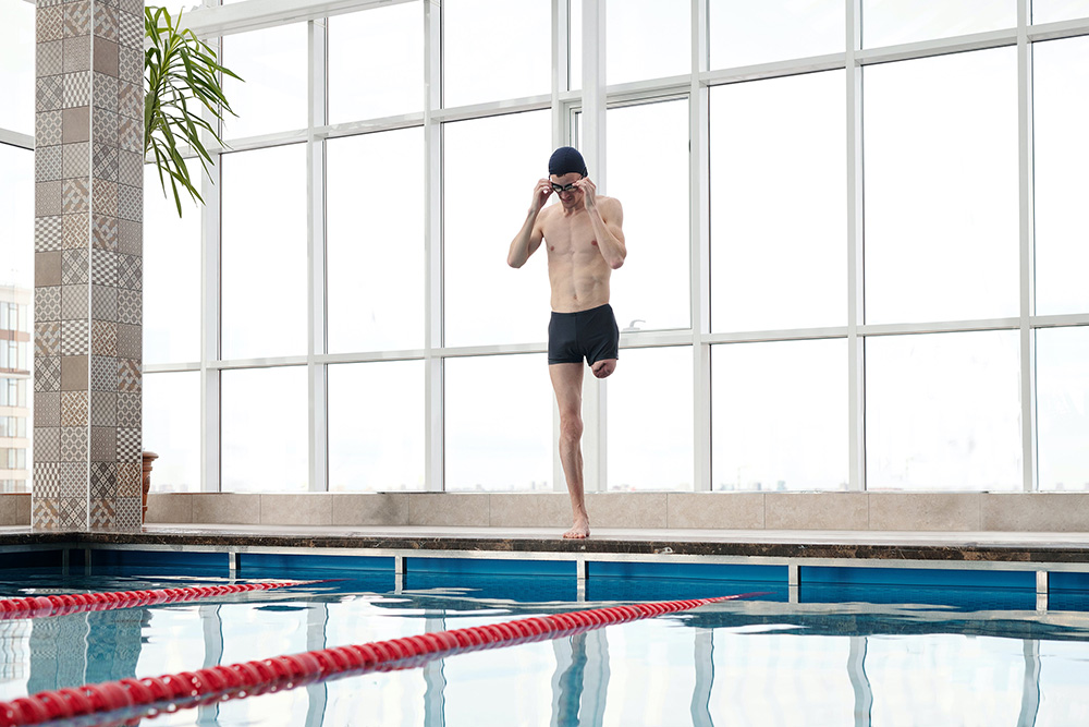 Ein Mann mit einer Behinderung steht am Rand eines Schwimmbeckens und setzt sich seine Schwimmbrille auf. Er trägt eine Badehose und eine Badekappe und bereitet sich darauf vor, ins Wasser zu springen. Dieses Bild symbolisiert Inklusion und zeigt, dass Menschen mit einer körperlichen Einschränkung ihre Ziele aktiv verfolgen können. Es verdeutlicht die Bedeutung von Unfallhilfe und Unterstützung nach einem Unfall, um Betroffenen zu helfen, ihre Lebensqualität zurückzugewinnen. Die Aufgabe des Unfallopfer-Hilfswerks ist es, Wege aufzuzeigen und Menschen mit Behinderung dabei zu unterstützen, ihre Unabhängigkeit und Mobilität zu erhalten.