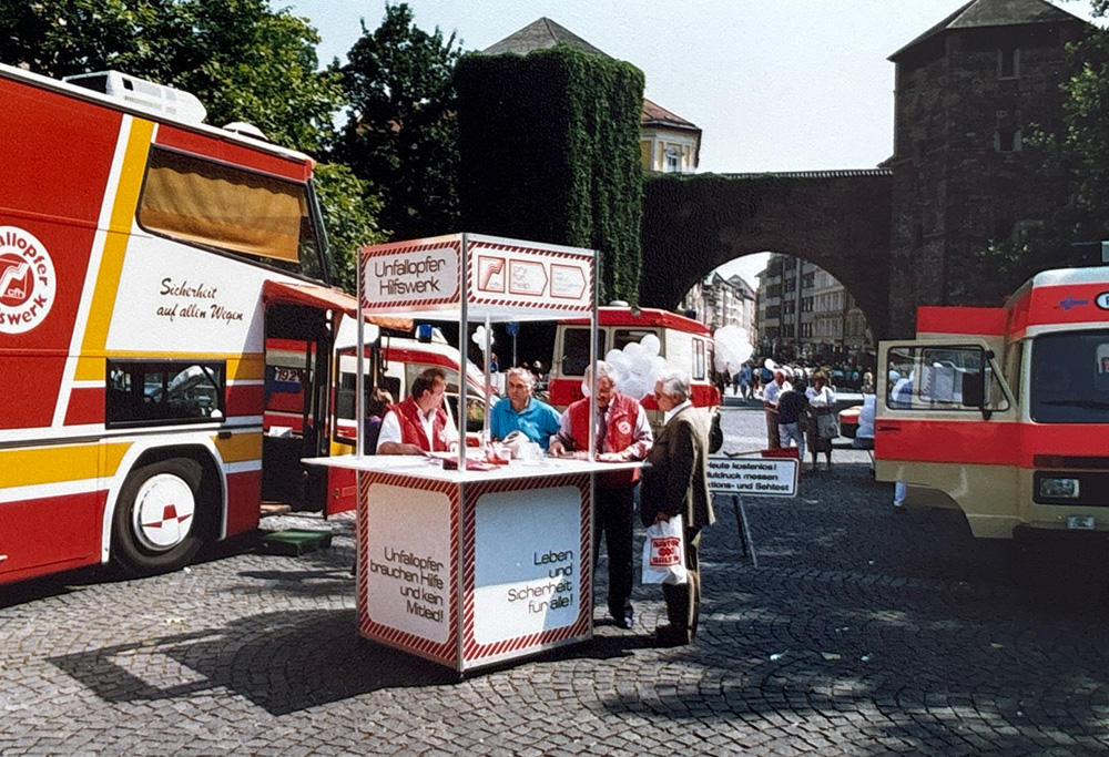 Ein Infostand des Unfallopfer-Hilfswerks steht in einer belebten Fußgängerzone. Menschen versammeln sich um den Stand, lassen sich beraten und informieren sich über Unterstützungsmöglichkeiten nach einem Unfall. Das Team des Unfallopfer-Hilfswerks steht bereit, um Fragen zu beantworten und Hilfsangebote vorzustellen. Die Szene symbolisiert direkte Hilfe, Aufklärung und die enge Verbindung zur Gemeinschaft, um Unfallopfern die bestmögliche Unterstützung zu bieten.