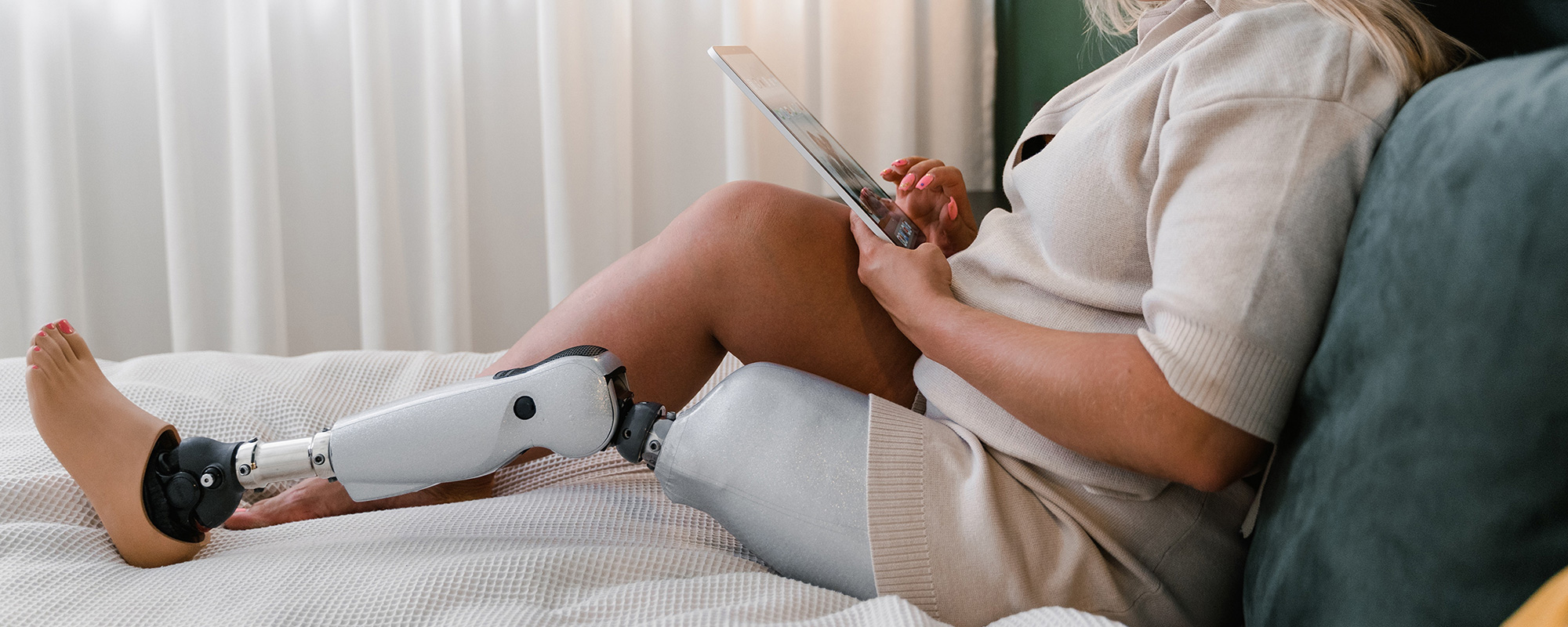 Eine Frau mit Beinprothese sitzt entspannt auf ihrem Bett und durchstöbert auf ihrem iPad den Onlineshop des Unfallopfer-Hilfswerks. Sie sucht nach gebrauchten Hilfsmitteln und innovativen Alltagshelfern, die ihren Alltag erleichtern. Das Bild symbolisiert die barrierefreie E-Commerce-Plattform, die Unfallopfern und Menschen mit Behinderung praktische Lösungen bietet – von Mobilitätshilfen bis zu spezialisierten Produkten für mehr Selbstständigkeit.