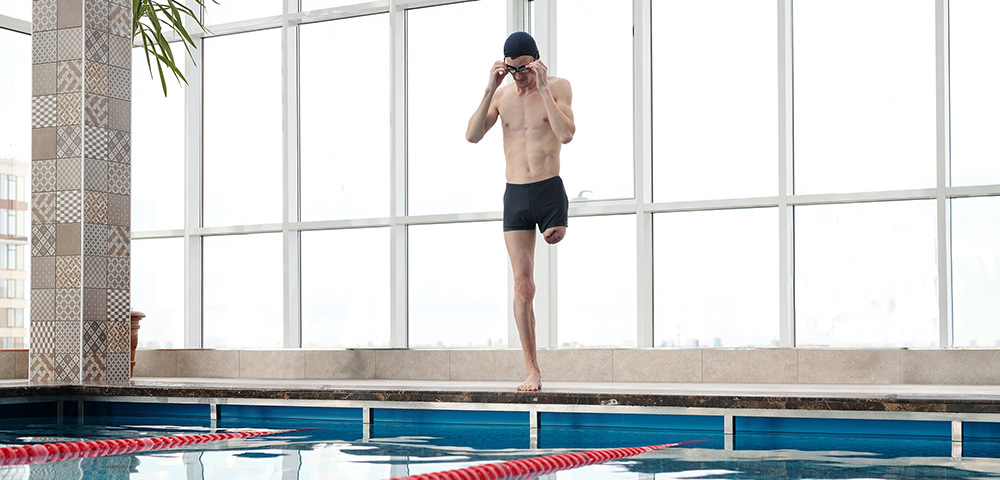 Ein Mann mit einer Behinderung steht am Rand eines Schwimmbeckens und setzt sich seine Schwimmbrille auf. Er trägt eine Badehose und eine Badekappe und bereitet sich darauf vor, ins Wasser zu springen. Dieses Bild symbolisiert Inklusion und zeigt, dass Menschen mit einer körperlichen Einschränkung ihre Ziele aktiv verfolgen können. Es verdeutlicht die Bedeutung von Unfallhilfe und Unterstützung nach einem Unfall, um Betroffenen zu helfen, ihre Lebensqualität zurückzugewinnen. Die Aufgabe des Unfallopfer-Hilfswerks ist es, Wege aufzuzeigen und Menschen mit Behinderung dabei zu unterstützen, ihre Unabhängigkeit und Mobilität zu erhalten.