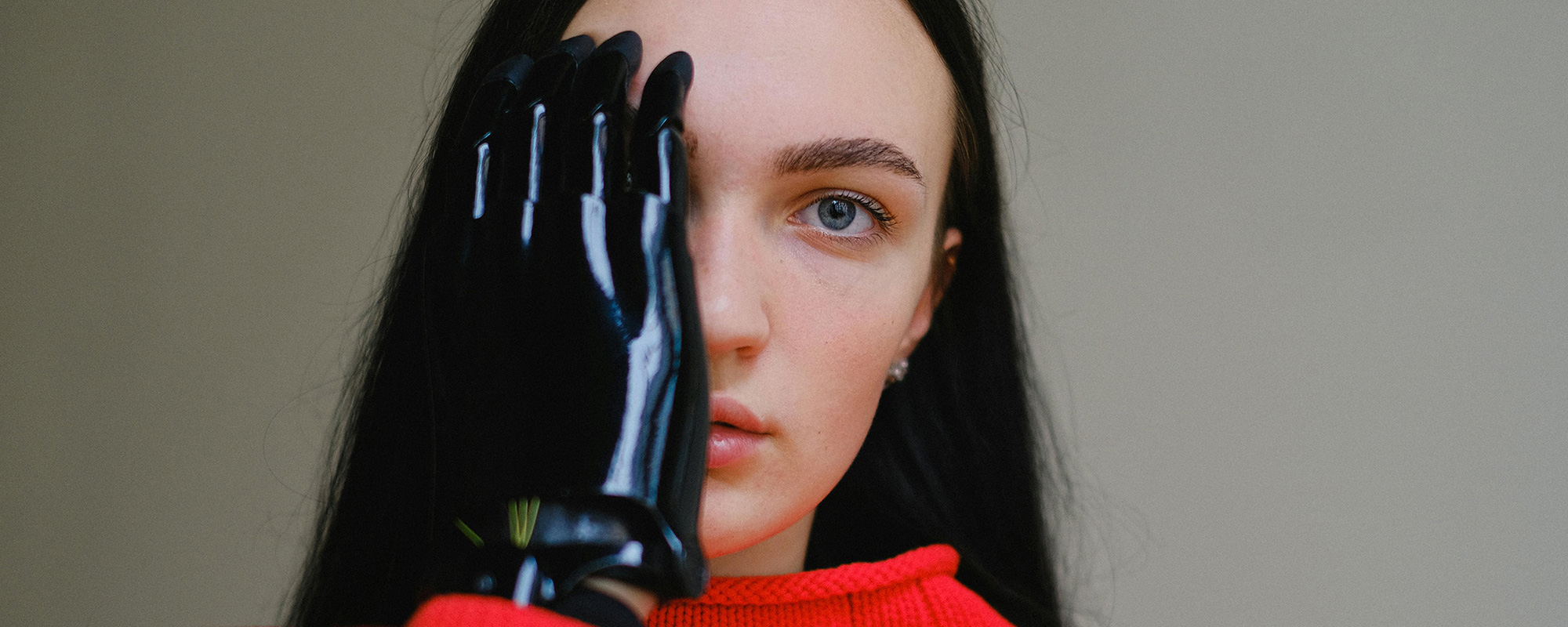 Kündigung - Unfallopfer Mitgliedschaft. Eine junge Frau mit schwarzen Haaren und rotem Pullover hält ihre Handprothese halb vor ihr Gesicht. Ihr verzweifelter Blick richtet sich direkt in die Kamera. Das Bild verdeutlicht, wie sehr Menschen auf die Unterstützung des Unfallopfer-Hilfswerks angewiesen sind. Es symbolisiert, dass jede Mitgliedschaft zählt, um Unfallopfern die nötige Hilfe und Versorgung zu ermöglichen.